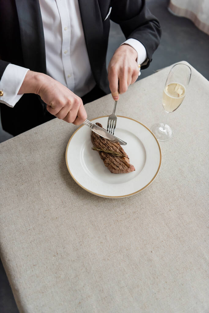 κορυφαία άποψη του πλούσιου άνδρα με σμόκιν κοπή νόστιμα μπριζόλα βοείου κρέατος στο πιάτο κοντά σε ένα ποτήρι σαμπάνια - Φωτογραφία, εικόνα