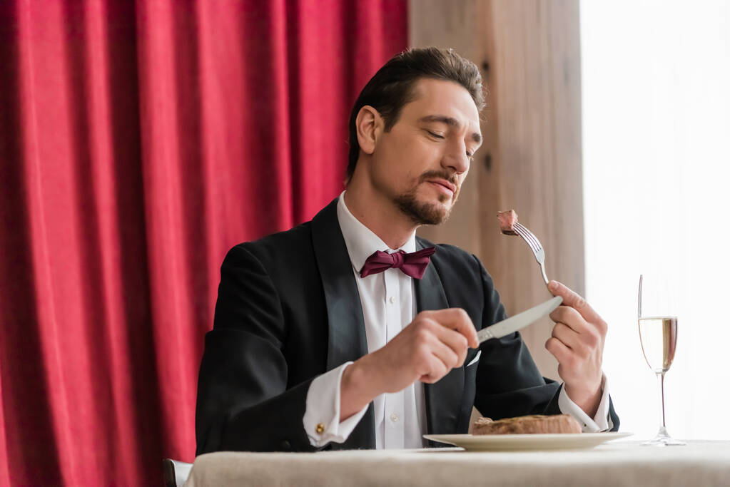 хорошо одетый мужчина в смокинге наслаждается вкусом говяжьего стейка возле шампанского в стакане на обеденном столе - Фото, изображение