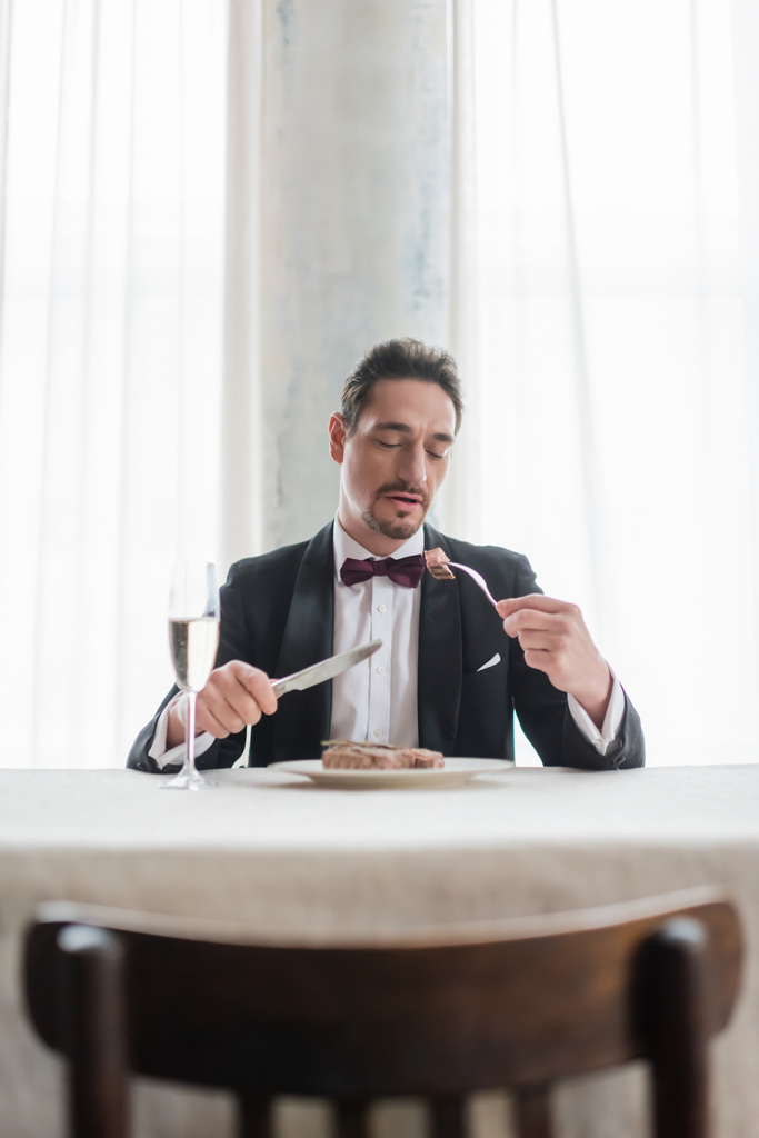 dobrze ubrany dżentelmen w smokingu jedzący pyszny stek wołowy przy kieliszku szampana, zamożne życie - Zdjęcie, obraz