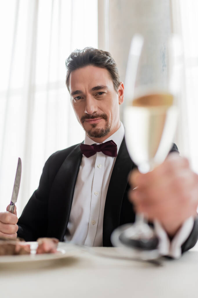 богатый человек в смокинге, приветствуя бокал шампанского рядом с вкусным бифштексом и глядя в камеру - Фото, изображение