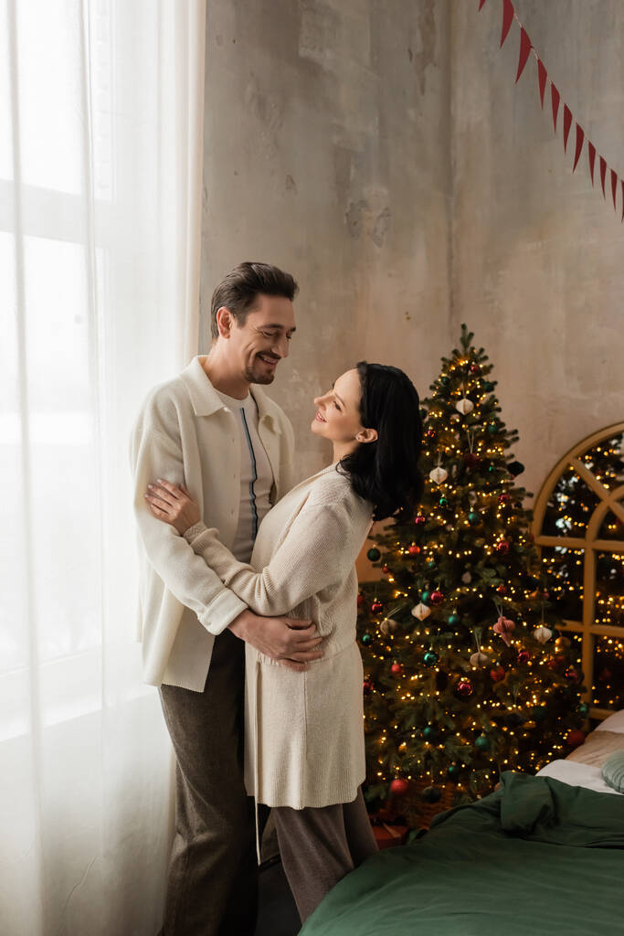 счастливая пара в удобной домашней одежде улыбается и обнимается рядом с украшенной елкой в спальне - Фото, изображение