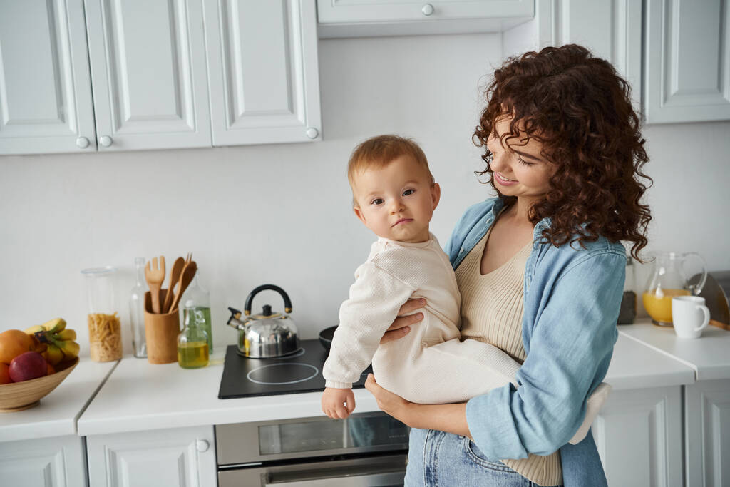 sonriente rizado mujer sosteniendo bebé niña en romperse mirando la cámara en acogedora cocina, familia feliz - Foto, imagen