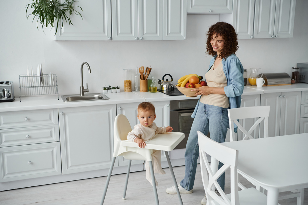 mujer sonriente con tazón de frutas frescas cerca del niño en silla de bebé en la cocina moderna, hora de la comida por la mañana - Foto, Imagen
