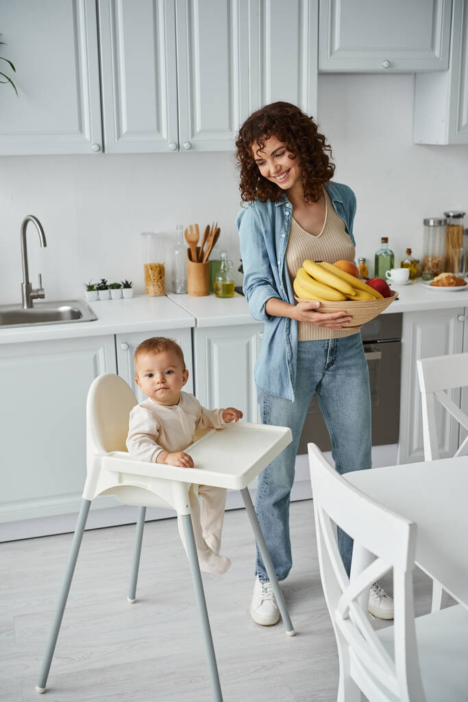 radosna kobieta trzymająca miskę świeżych owoców w pobliżu dziecka w foteliku dziecięcym we współczesnej kuchni, śniadanie - Zdjęcie, obraz