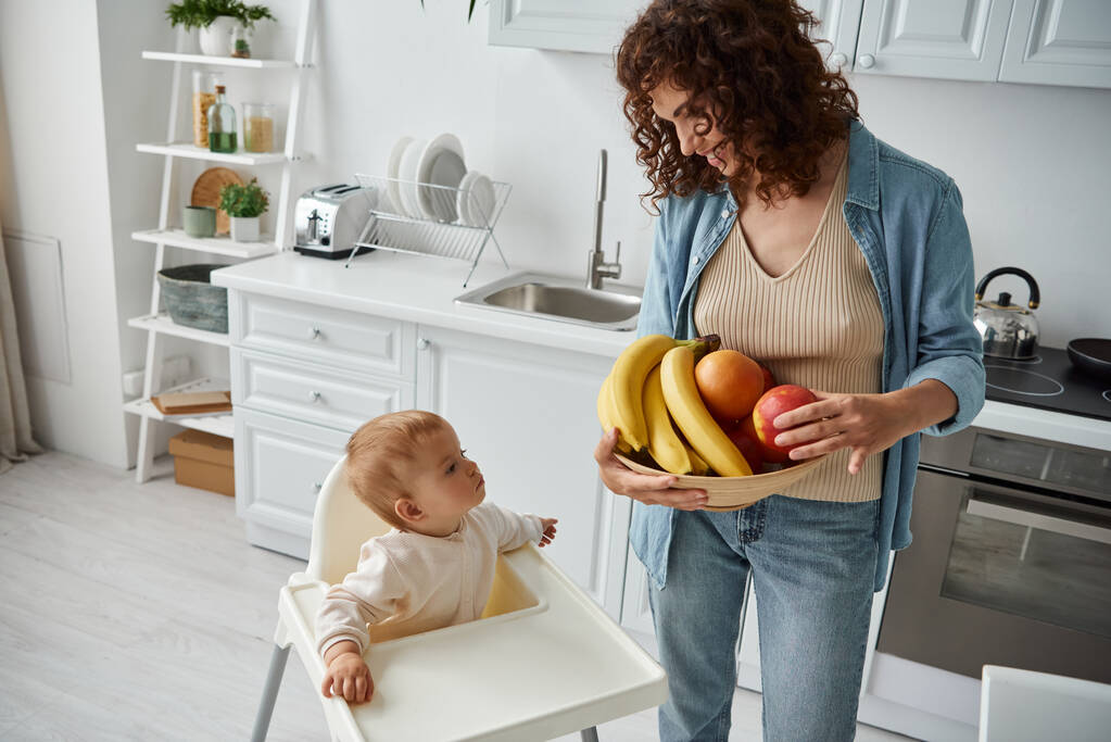 mujer feliz con tazón de plátanos y manzanas cerca de la pequeña hija sentada g en silla de bebé en la cocina - Foto, Imagen