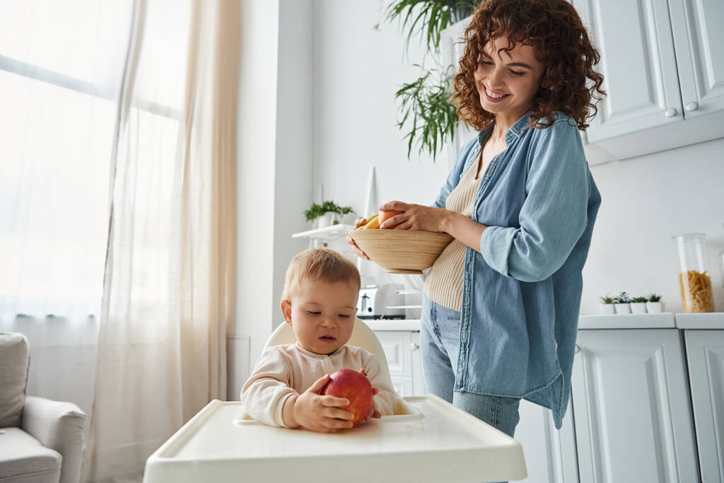 mujer alegre con tazón de frutas frescas mirando a la hija del niño sosteniendo manzana madura en silla de bebé - Foto, Imagen