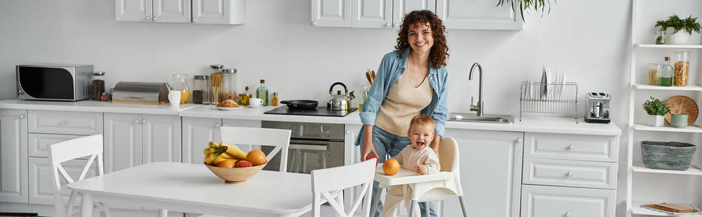 взволнованная мать и дочь малыша, глядя на камеру рядом со свежими фруктами на современной кухне, баннер - Фото, изображение