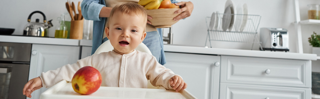 criança alegre na cadeira do bebê olhando maçã madura perto da mãe com tigela de frutas frescas, banner - Foto, Imagem