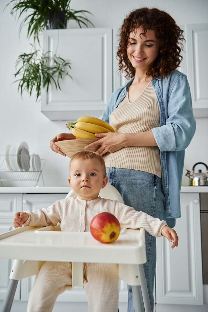 χαμογελαστή γυναίκα κρατώντας μπολ με φρέσκα φρούτα κοντά στην κόρη νήπιο σε καρέκλα μωρού με ώριμο μήλο - Φωτογραφία, εικόνα