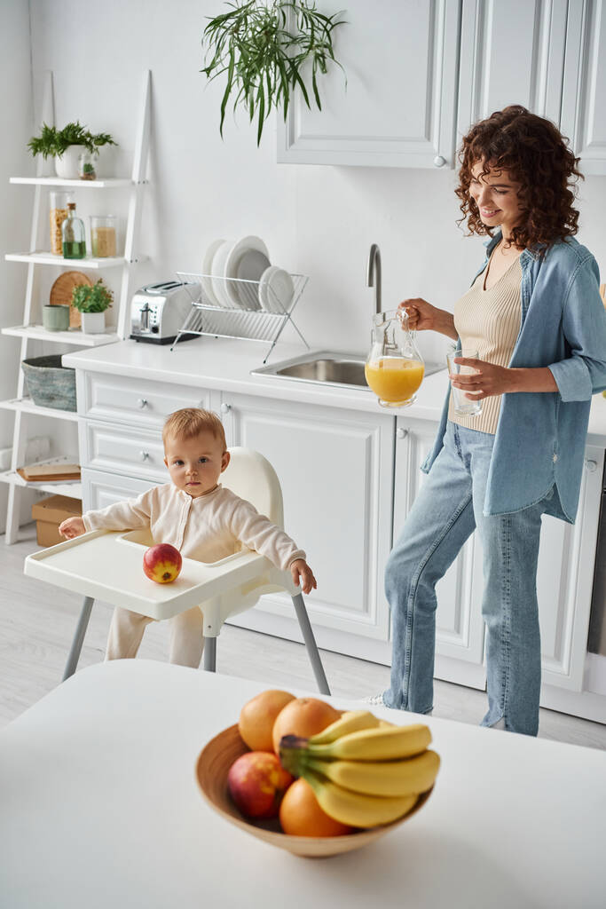gelukkig vrouw met kruik van verse sinaasappelsap in de buurt van kind in kinderstoel en rijp fruit in moderne keuken - Foto, afbeelding
