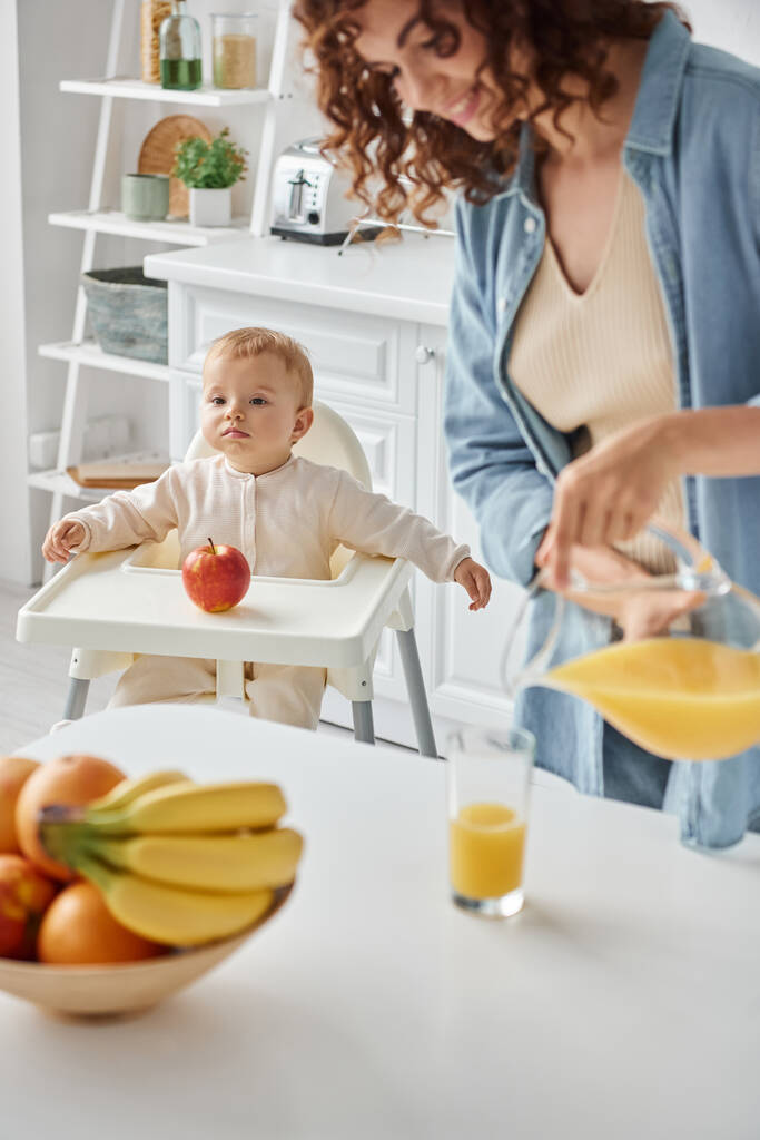 lindo niño sentado en silla de bebé cerca de manzana mientras mamá feliz verter jugo de naranja fresca para el desayuno - Foto, imagen