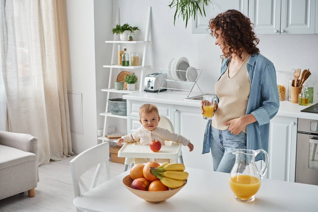 mujer rizada con vaso de jugo de naranja mirando al niño en silla de bebé en la cocina con frutas maduras - Foto, Imagen