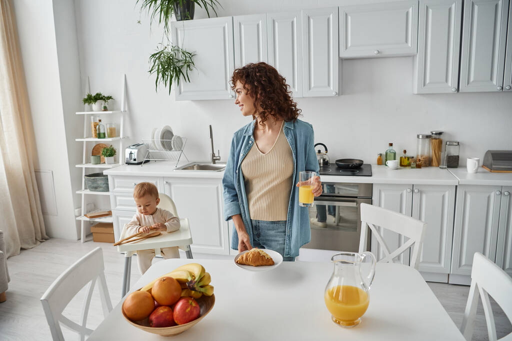 mulher com suco de laranja e croissant perto de frutas e criança brincando com pinças de madeira na cozinha - Foto, Imagem