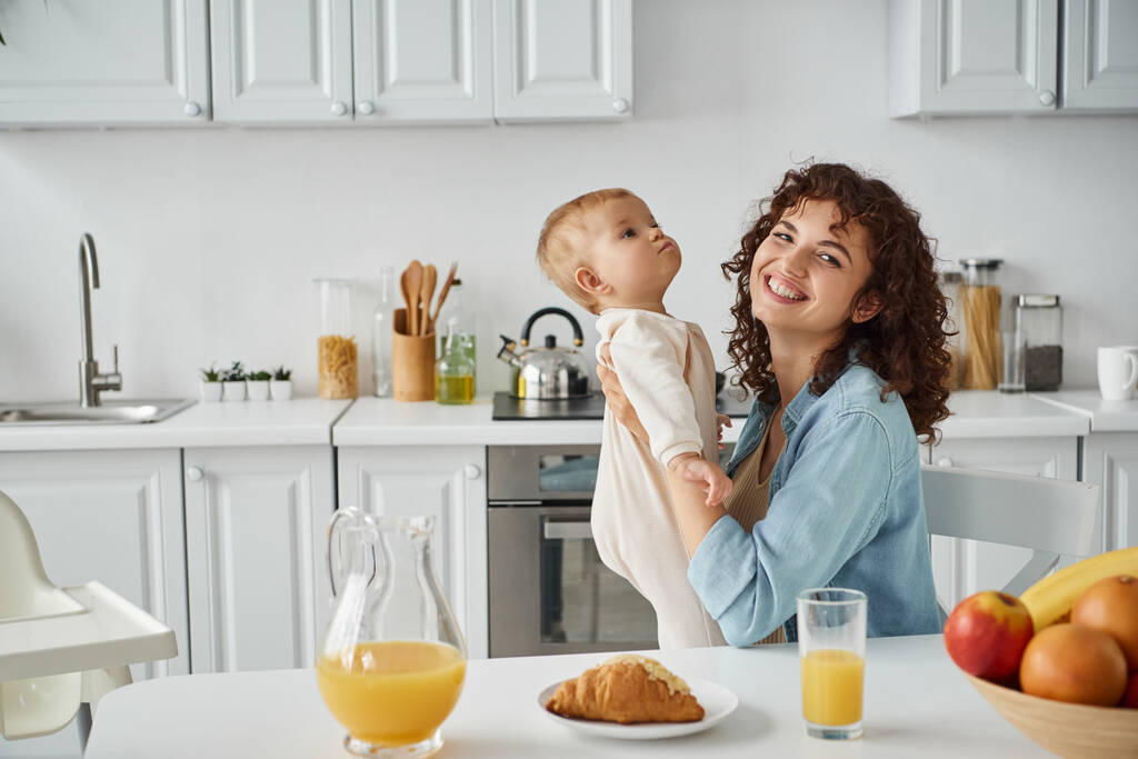 freudige Mutter hält Baby-Mädchen in der Nähe von Croissant und Orangensaft mit Früchten in der Küche, fröhliches Frühstück - Foto, Bild