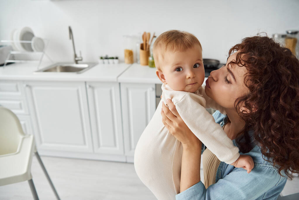 mujer rizada abrazando y besando a la hija pequeña en mamada en la acogedora cocina en casa, felicidad familiar - Foto, imagen