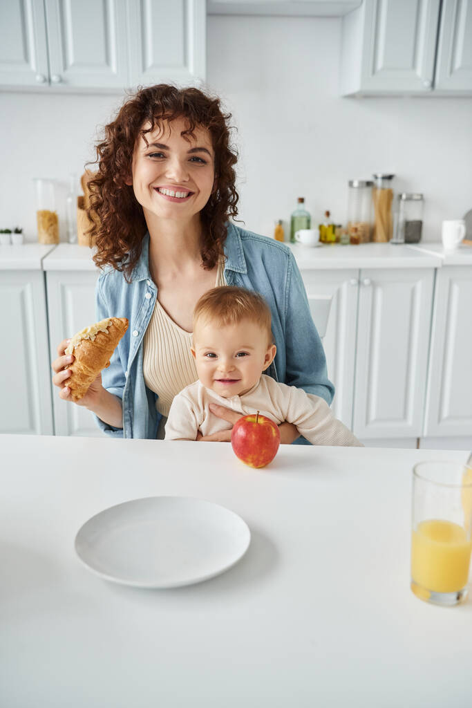 matka a batole dítě s úsměvem v blízkosti croissant a čerstvý pomerančový džus s jablkem při snídani - Fotografie, Obrázek
