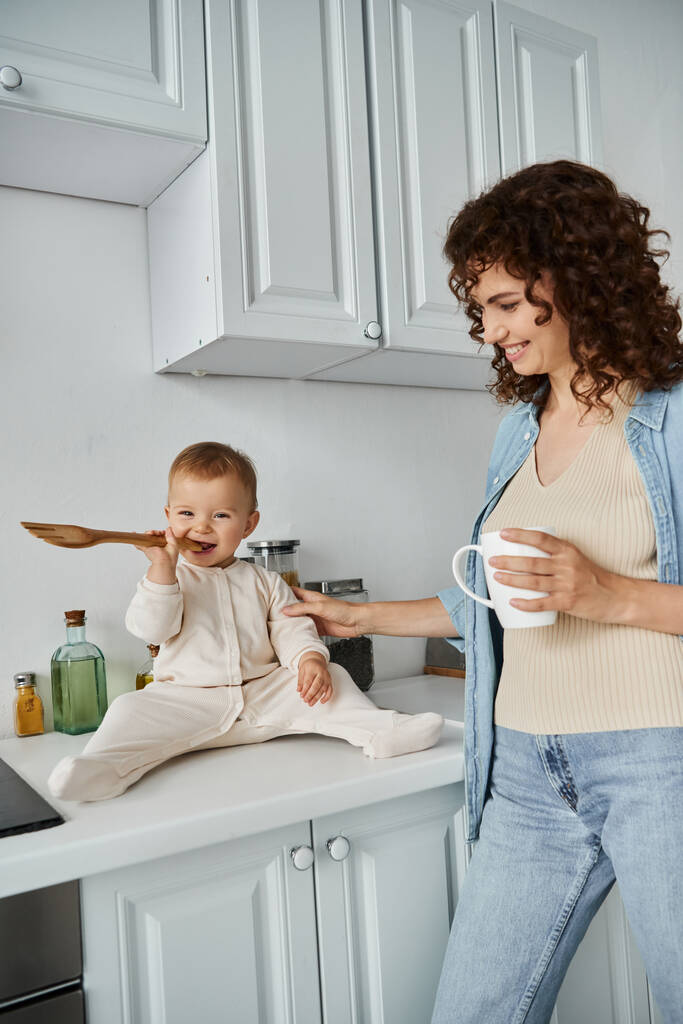 carefree baby sitting on kitchen counter and chewing wooden fork near smiling mother with coffee cup - Photo, Image