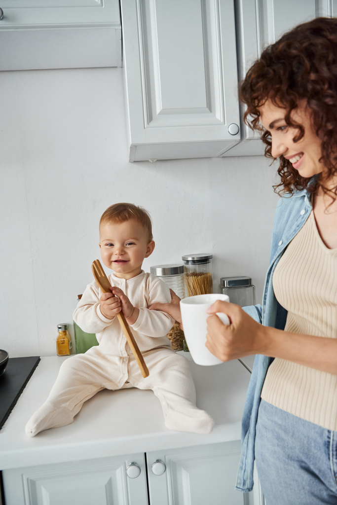 веселый ребенок сидит на кухонном столе и играет с деревянной вилкой рядом счастливая мама с чашкой кофе - Фото, изображение
