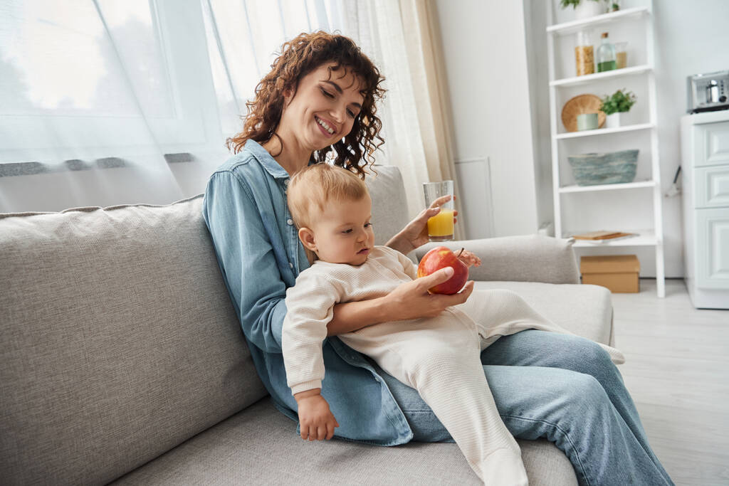 joyful woman holding apple and orange juice while sitting on cozy couch with toddler daughter - Photo, Image