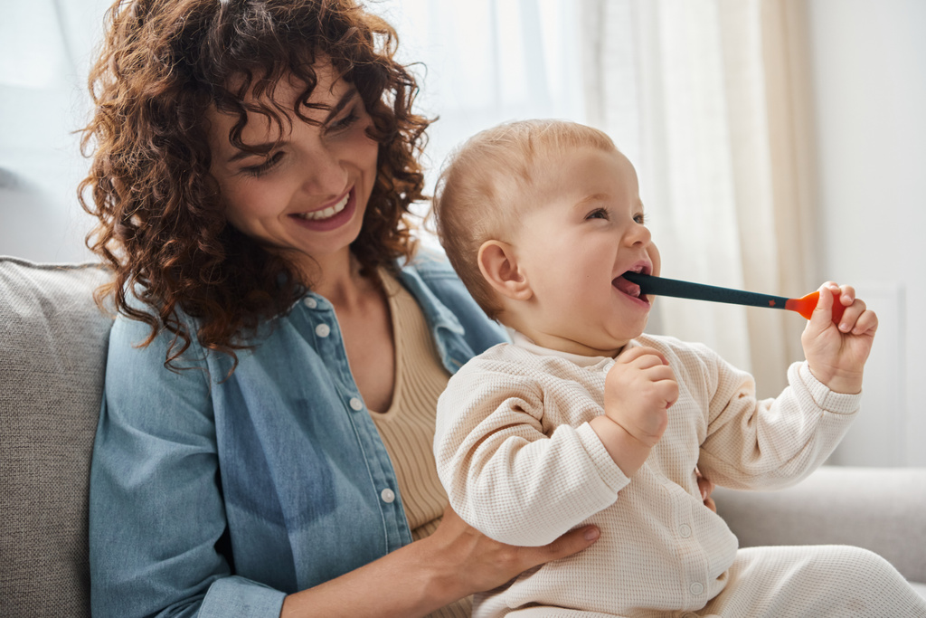 mujer sonriente sentada en el sofá con la hija pequeña masticando el juguete de dentición en la sala de estar, tiempo de juego - Foto, Imagen