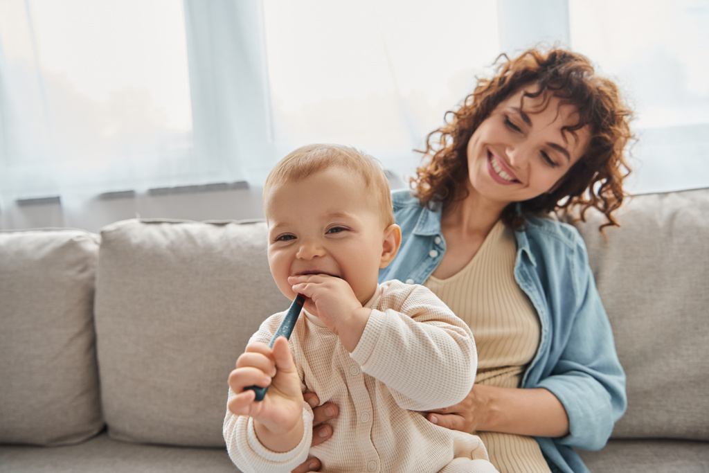 leikkisä vauva pureskelu kasvukipuja lelu lähellä hymyilevä äiti sohvalla olohuoneessa, laatuaikaa kotona - Valokuva, kuva