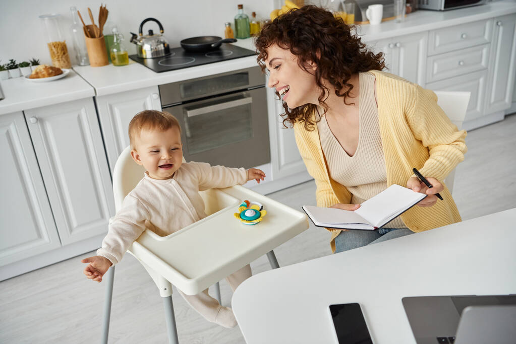 χαρούμενη μητέρα με σημειωματάριο κοντά ενθουσιασμένοι παιδί κάθεται σε καρέκλα μωρού με κουδουνίστρα παιχνίδι, γονέας εργασίας - Φωτογραφία, εικόνα