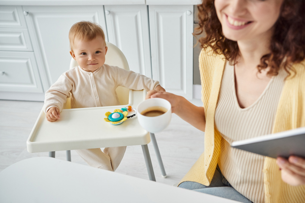 mujer con café de la mañana y portátil cerca de niño sonriente en silla de bebé con juguete sonajero en la cocina - Foto, Imagen