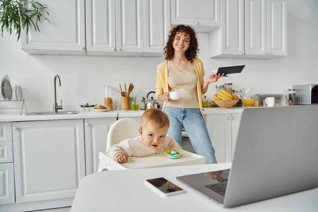 gelukkig vrouwelijke freelancer met koffiebeker en notebook in de buurt van kind in kinderstoel en apparaten in de keuken - Foto, afbeelding