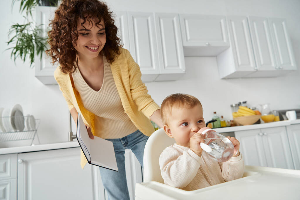 fröhliche Mutter mit Notizbuch blickt auf Kleinkind Tochter trinkt aus Babyflasche in Küche - Foto, Bild