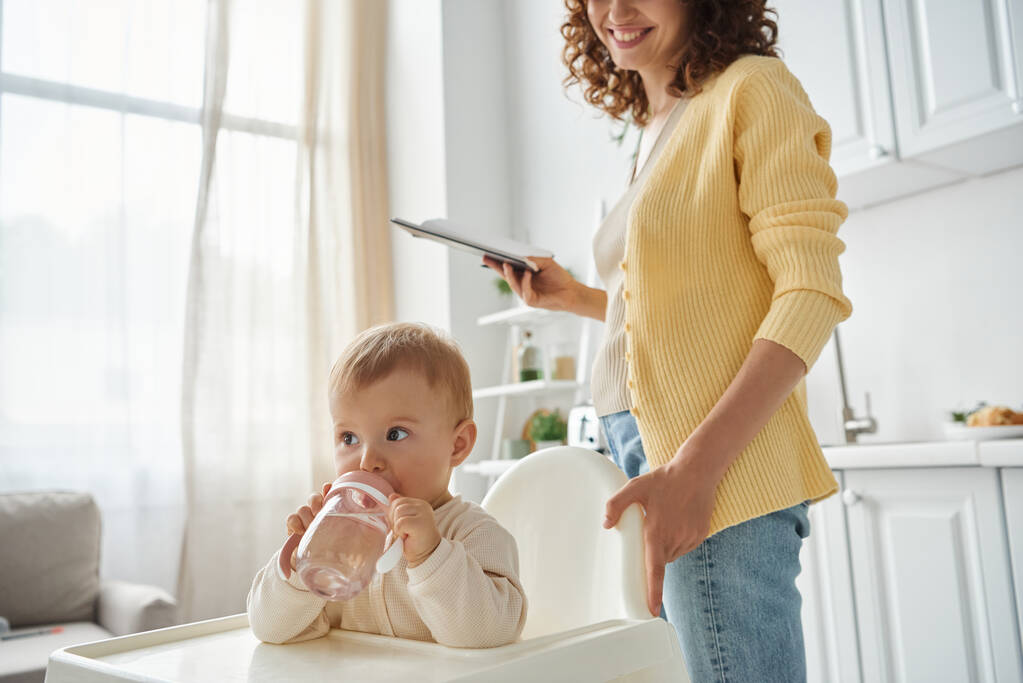 klein meisje zitten in baby stoel en het drinken van water in de buurt van glimlachende moeder met notebook in de keuken - Foto, afbeelding