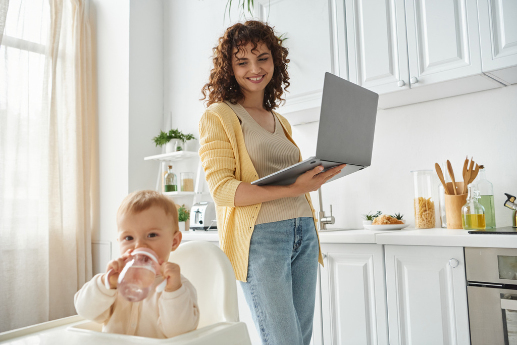 mujer feliz con el ordenador portátil mirando a la hija del niño bebiendo agua del biberón en la cocina - Foto, imagen