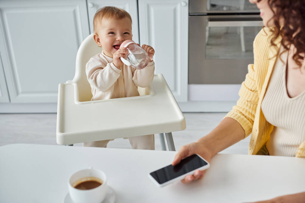 niño alegre bebiendo de biberón cerca de mamá con teléfono inteligente con pantalla en blanco cerca de la taza de café - Foto, Imagen