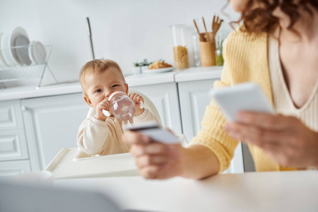 klein meisje drinken uit baby fles in de buurt van mam met smartphone en creditcard op wazig voorgrond - Foto, afbeelding