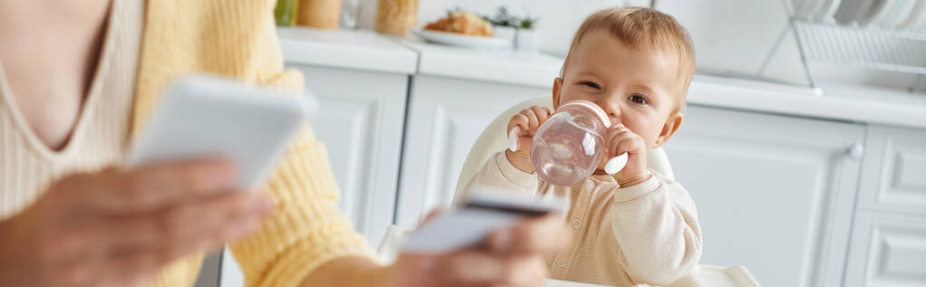 スマートフォンとクレジットカード,バナー付きのぼやけたママ近くの赤ちゃんボトルから飲む赤ちゃんの女の子 - 写真・画像
