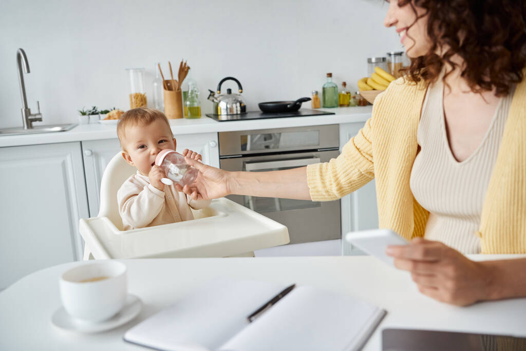 Frau mit Handy hilft kleinem Mädchen beim Trinken aus Babyflasche während der Arbeit in der Küche - Foto, Bild