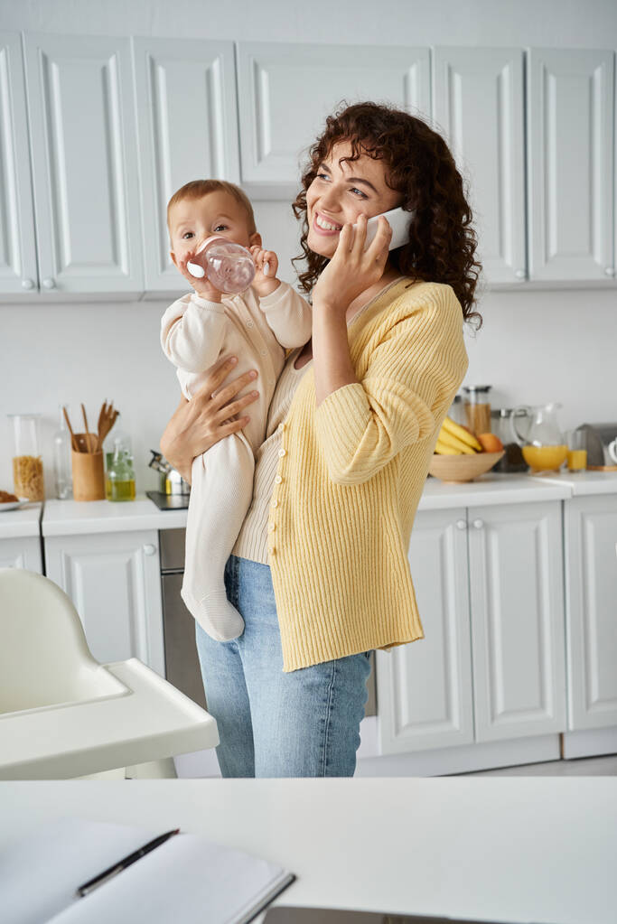 alegre madre hablando en smartphone y sosteniendo al niño bebiendo del biberón en la cocina - Foto, Imagen