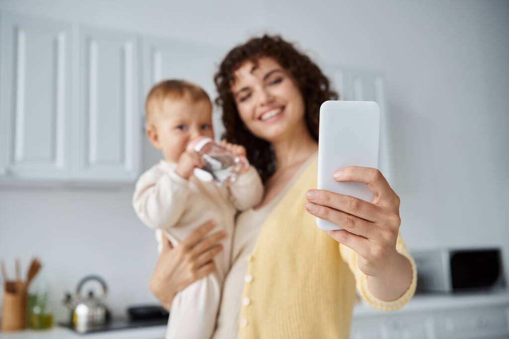 onnellinen äiti ottaa selfie älypuhelimeen vauvan tyttö juo vauvan pullo keittiössä - Valokuva, kuva
