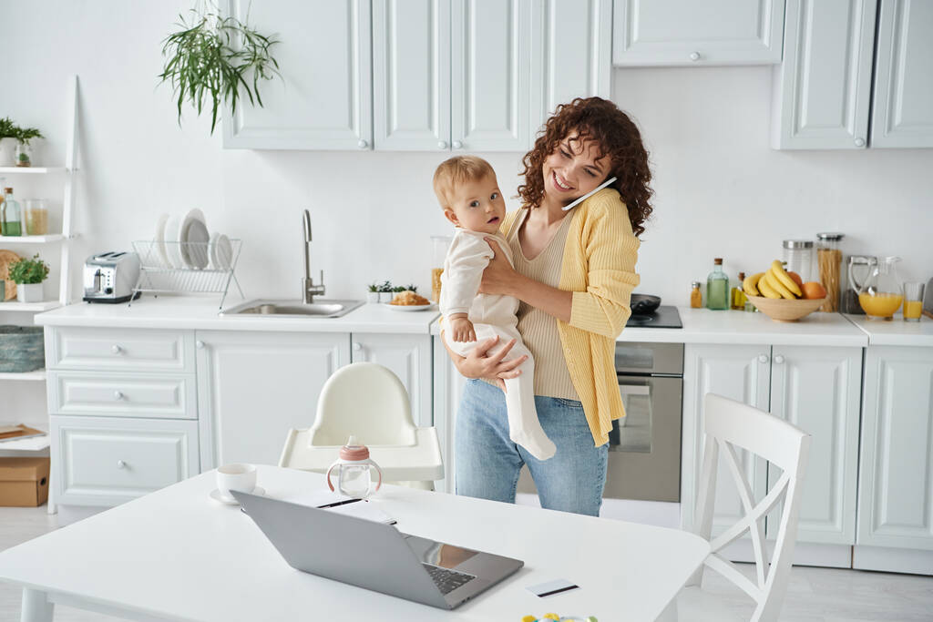 femme tenant bébé et parlant sur smartphone près d'un ordinateur portable dans la cuisine moderne, équilibre de la vie professionnelle - Photo, image