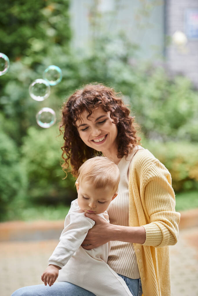retrato de mujer alegre con hija pequeña cerca de burbujas de jabón volando pasar tiempo al aire libre - Foto, Imagen
