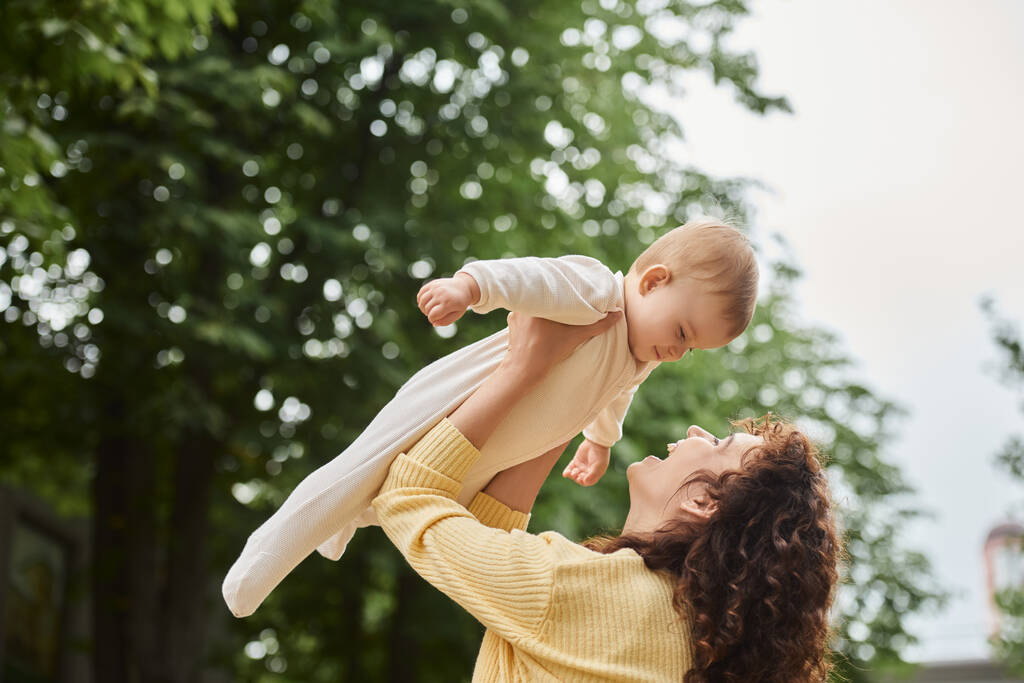 mulher animada levantando filhinha nas mãos enquanto se diverte ao ar livre, maternidade feliz - Foto, Imagem