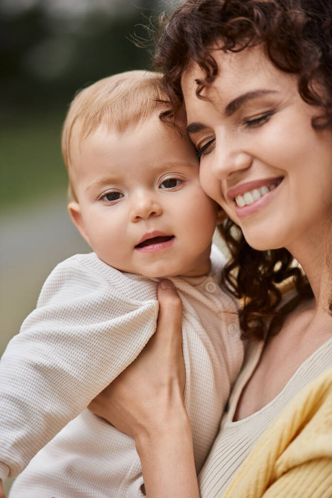 Porträt einer lächelnden Frau mit geschlossenen Augen, die ihr Kleinkind im Freien umarmt, glückliche Momente - Foto, Bild