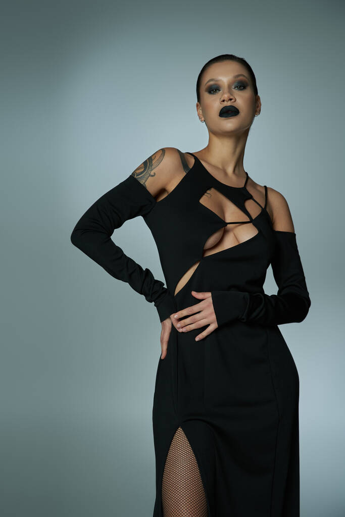 mujer tatuada con maquillaje oscuro espeluznante vestido de halloween negro y mirando a la cámara en gris - Foto, Imagen