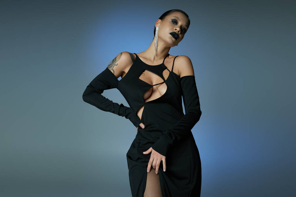 ďábelská krása, žena v tmavém make-upu a černé šaty pózují s rukou na boku na modrém šedém pozadí - Fotografie, Obrázek
