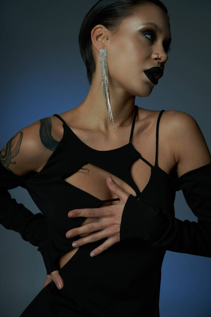 mulher tatuada em vestido elegante preto e maquiagem sinistra olhando para o fundo azul e cinza - Foto, Imagem