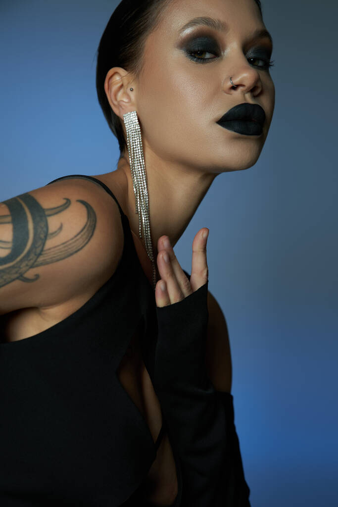 portrait of tattooed enchanting woman with dark makeup looking at camera on blue and grey backdrop - Photo, Image