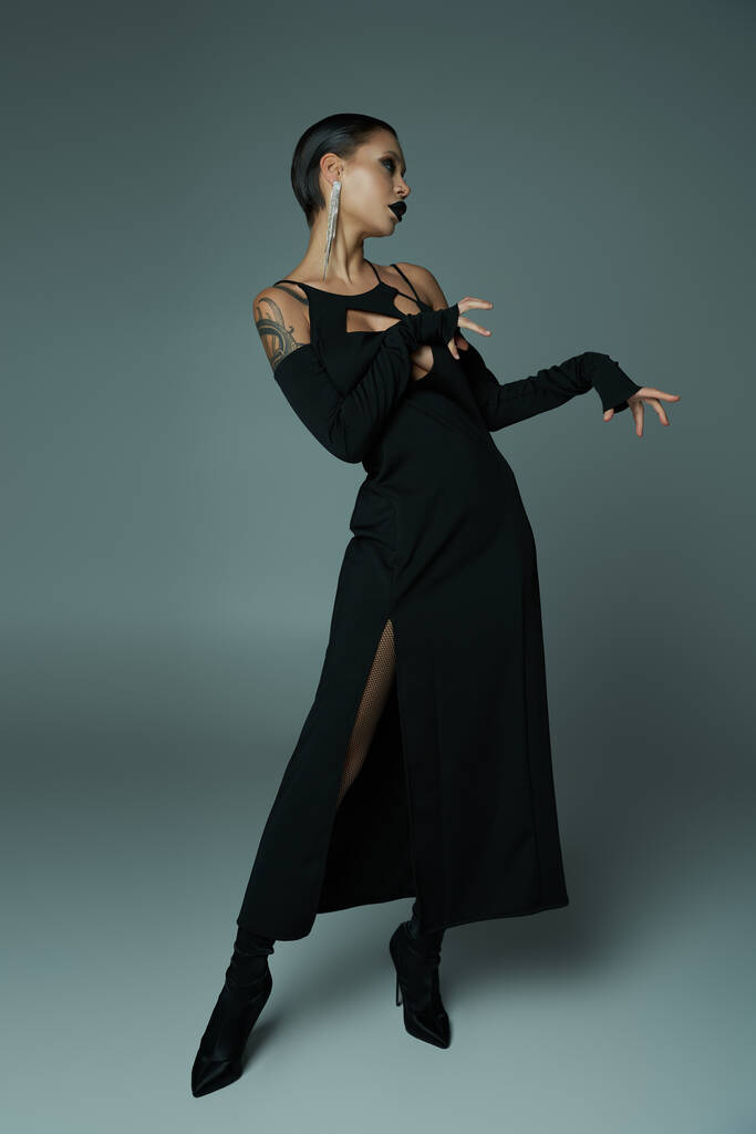 σκούρα ομορφιά, γυναίκα με τατουάζ σε μαύρο αποκριάτικο φόρεμα και απόκοσμο μακιγιάζ ποζάροντας σε γκρι, ολόσωμη - Φωτογραφία, εικόνα