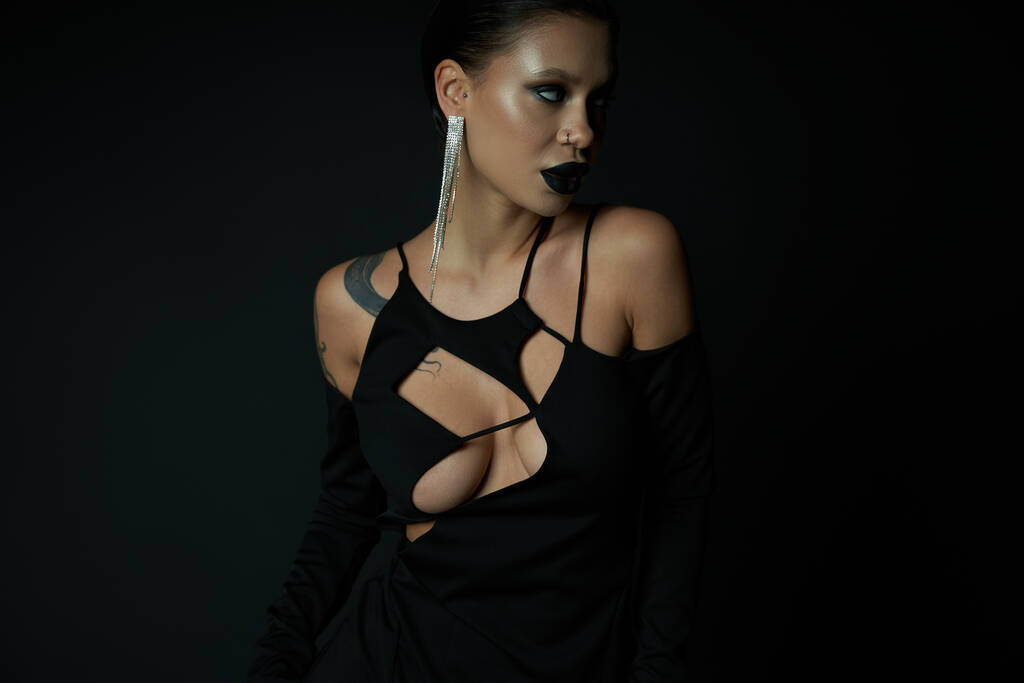 mujer tatuada en pendiente brillante y vestido de bruja oscura mirando hacia otro lado en negro, concepto de halloween - Foto, Imagen