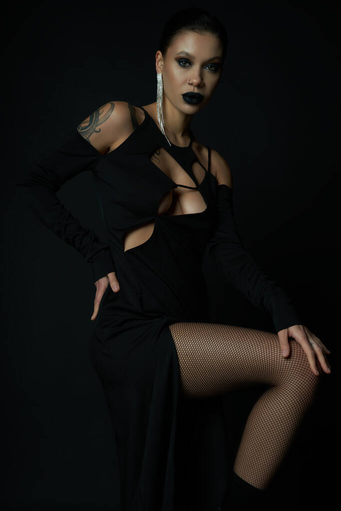 mujer tatuada con estilo en vestido de halloween negro y maquillaje espeluznante posando con la mano en la cadera en negro - Foto, Imagen