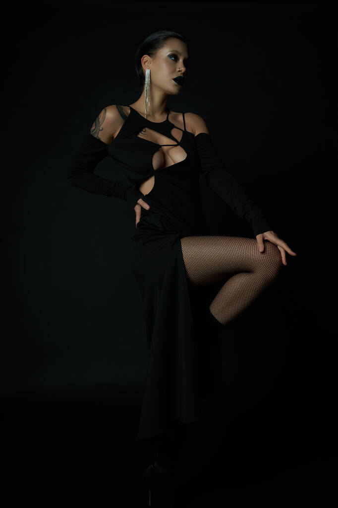 σαγηνευτική γυναίκα με τατουάζ σε μαύρο αποκριάτικο φόρεμα και σκούρο μακιγιάζ κοιτάζοντας μακριά, ενώ θέτουν σε μαύρο - Φωτογραφία, εικόνα