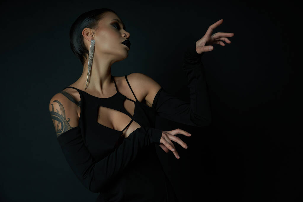 сексуальна татуйована жінка в гламурній сукні на Хеллоуїн і моторошний макіяж жестикулює на чорному тлі - Фото, зображення
